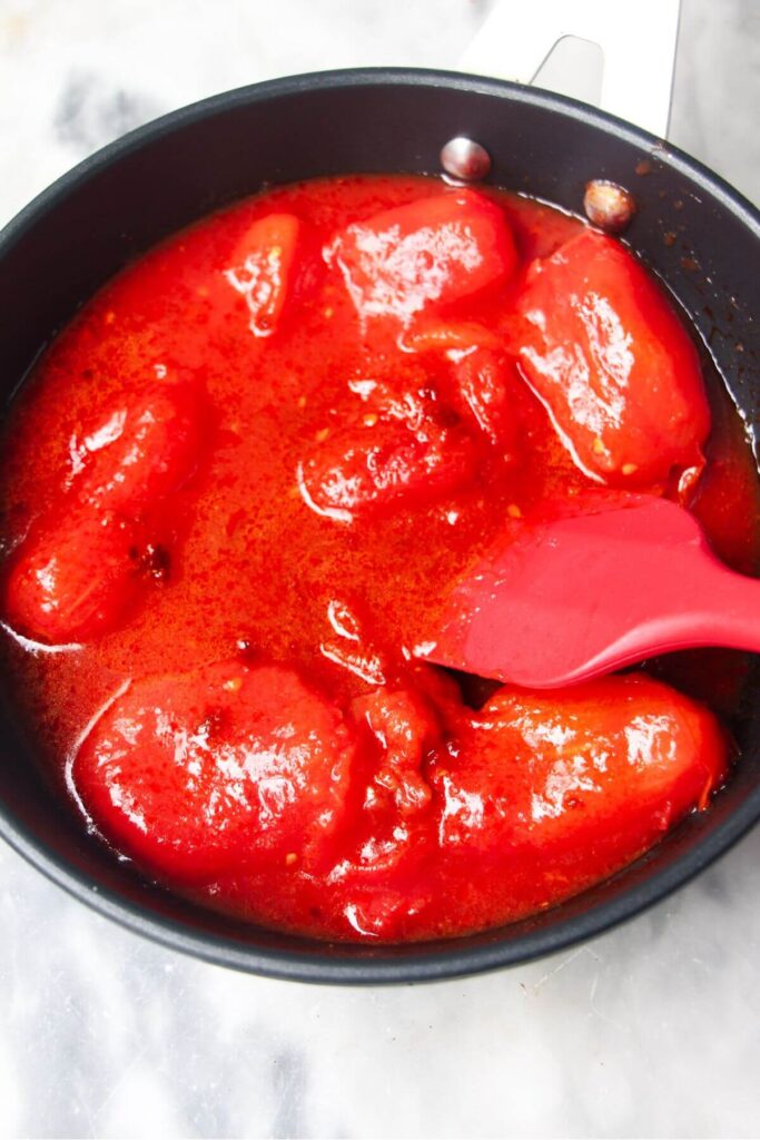 Tinned plum tomatoes added to a small black pan and being stirred by a red spatula.
