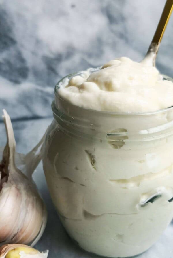 Vegan garlic aioli in glass jar with garlic bulb on side and spoon in the jar.