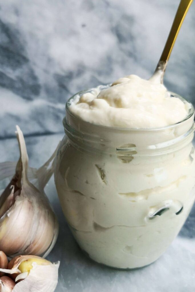 Vegan garlic aioli in a glass jar with a gold spoon inside, with garlic bulb on the side.