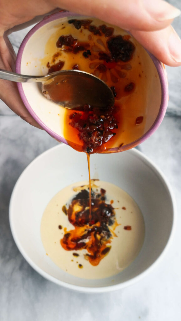 A small bowl of chilli oil being added to a bowl of tahini.