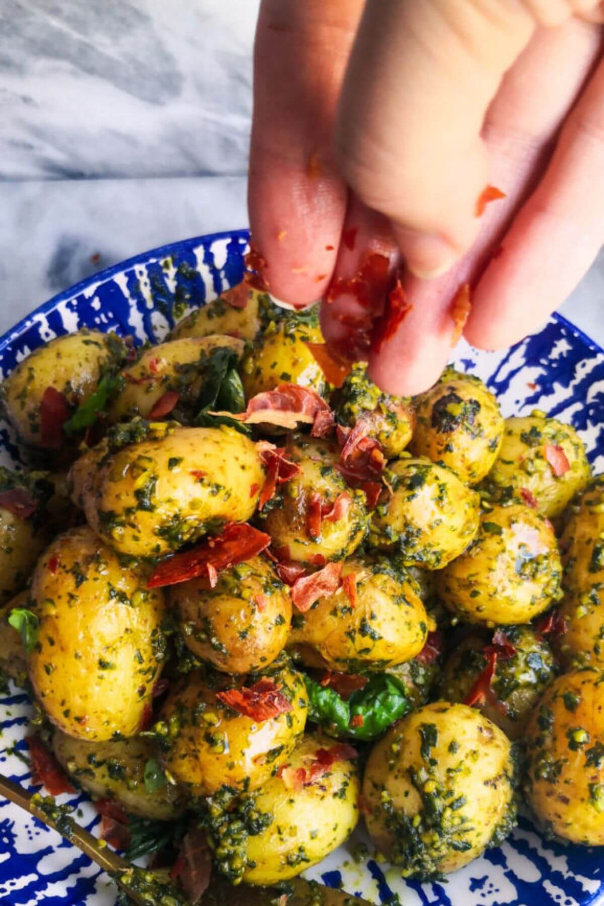 Hand sprinkling crispy prosciutto onto the pesto potatoes in a white and blue bowl.