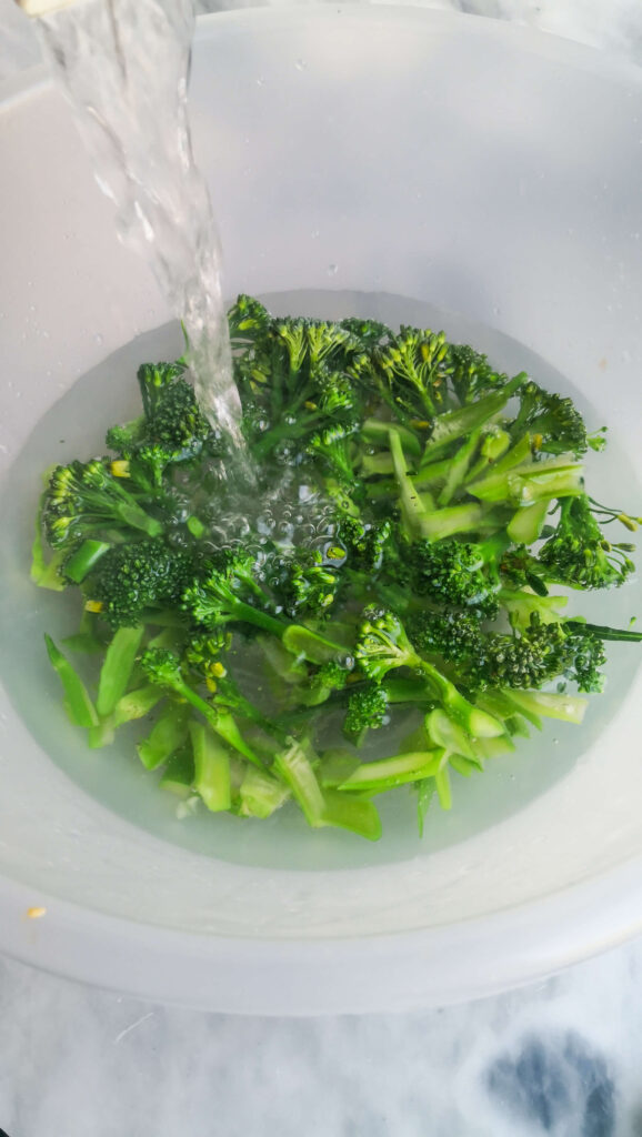 Boiling water being poured into a large bowl filled with sliced broccolini.