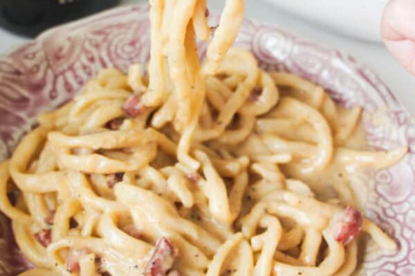 Fork holding up strands of aspaghetti, with a plate of carbonara in the foreground.