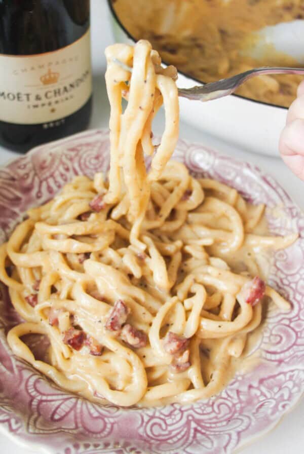 Fork holding up strands of aspaghetti, with a plate of carbonara in the foreground.