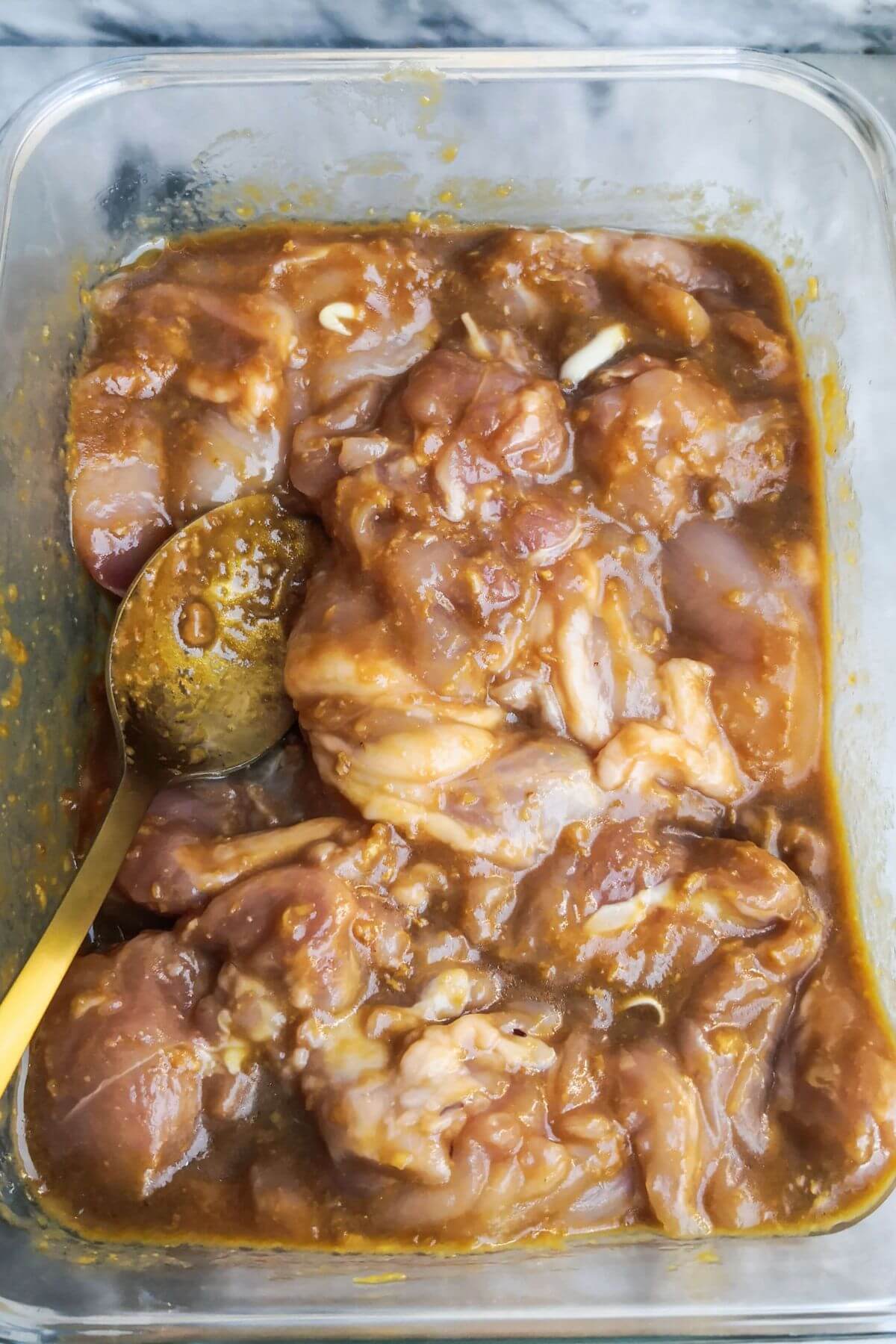 Chicken thighs mixed through marinade in a glass container with a gold spoon inside.