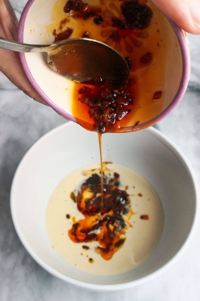 Chili oil being poured into a small bowl with tahini in it.