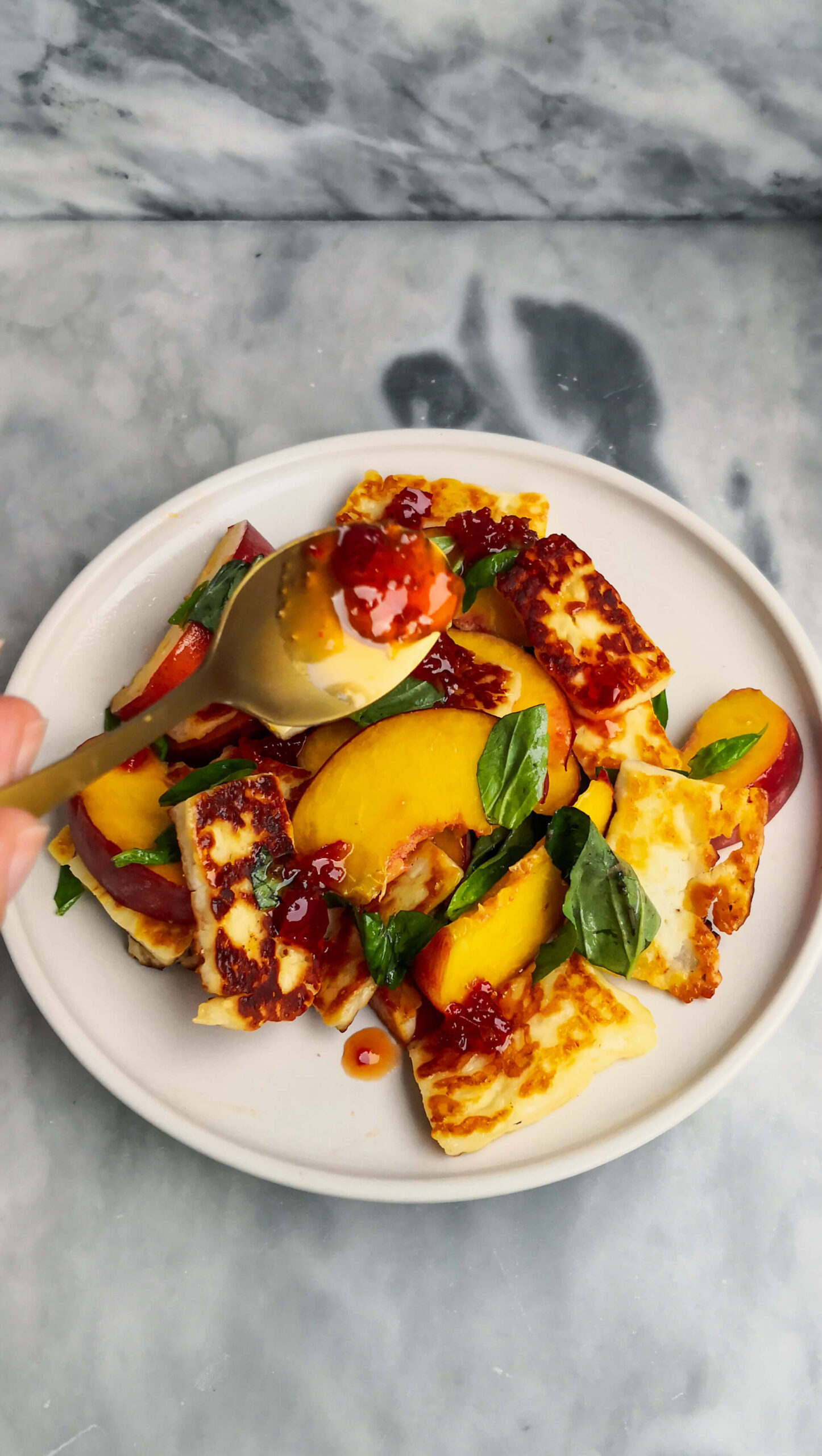 A small gold spoon drizzling chilli jam onto peach and halloumi salad on a grey marble background.