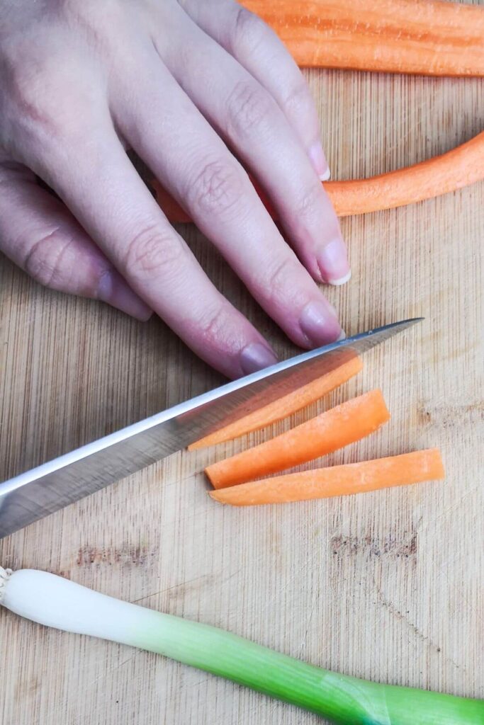 Chopping piece of carrot into thin strips with a large knife on a wooden background.
