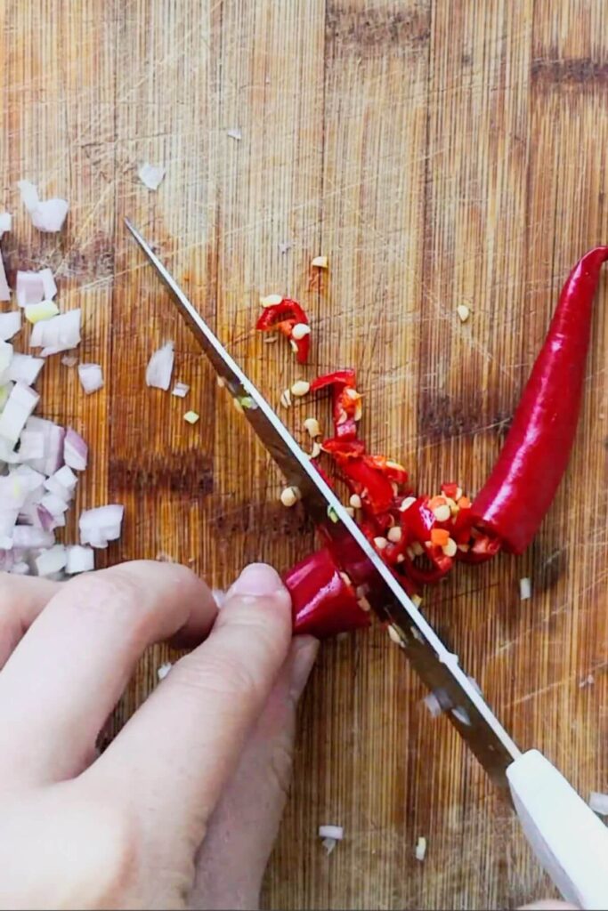 Hand with knife chopping red chilli on a wooden board.