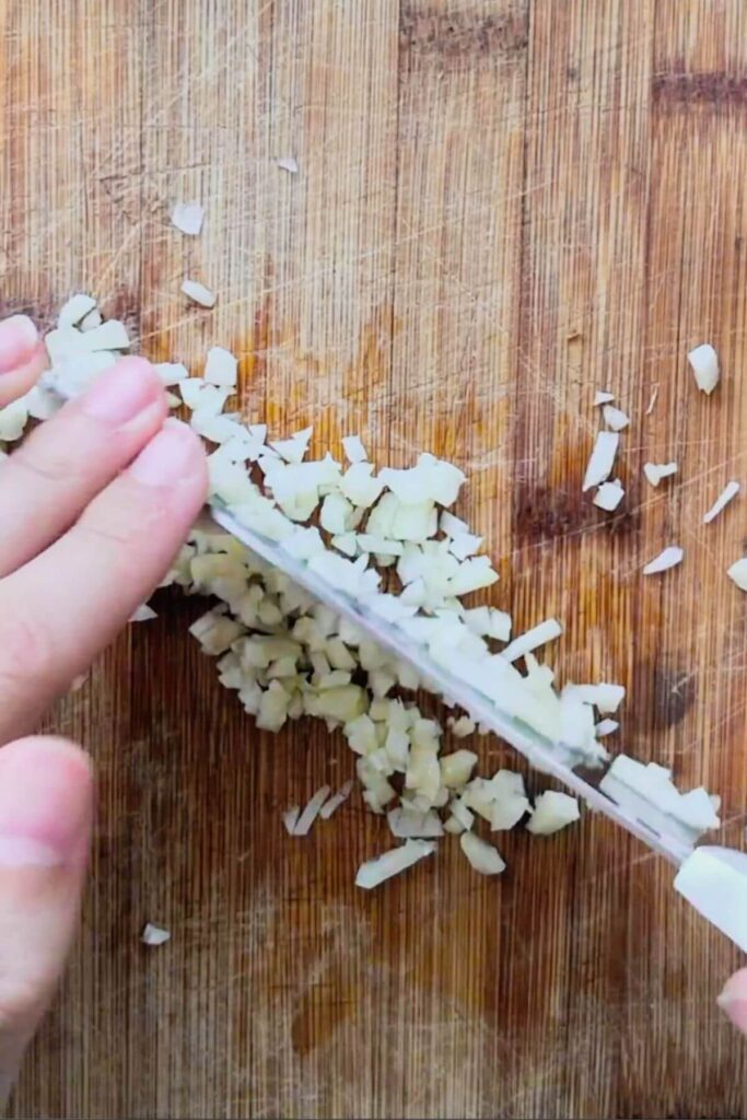 Hand with knife chopping garlic finely on a wooden board.