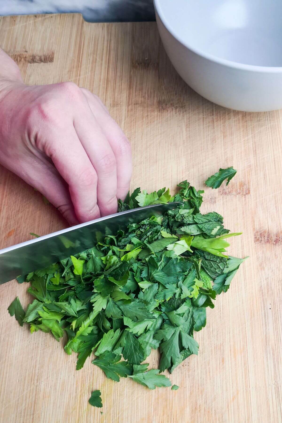 Finely chopping parsley leaves on a wooden board with a grey bowl in the background.