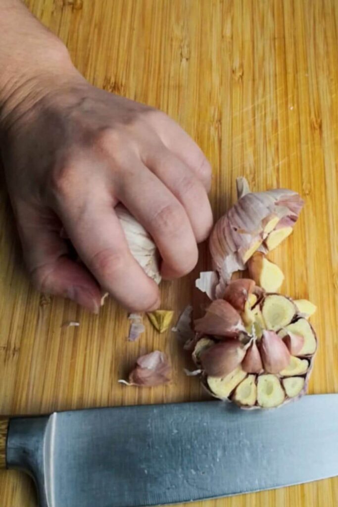 Whole head of garlic cut in half on a wooden board, with a hand holding the other side of the garlic.