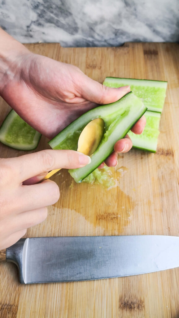 Hand holding a halved cucumber with a gold spoon scooping out the seeds.