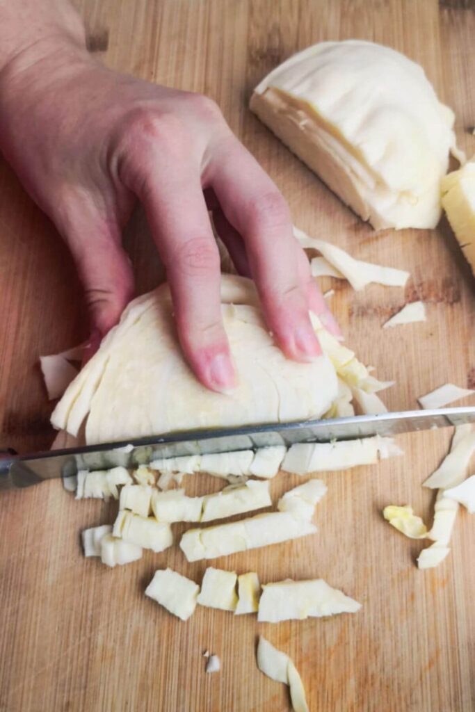 Chopping white cabbage into small cubes with a knife on a wooden board.