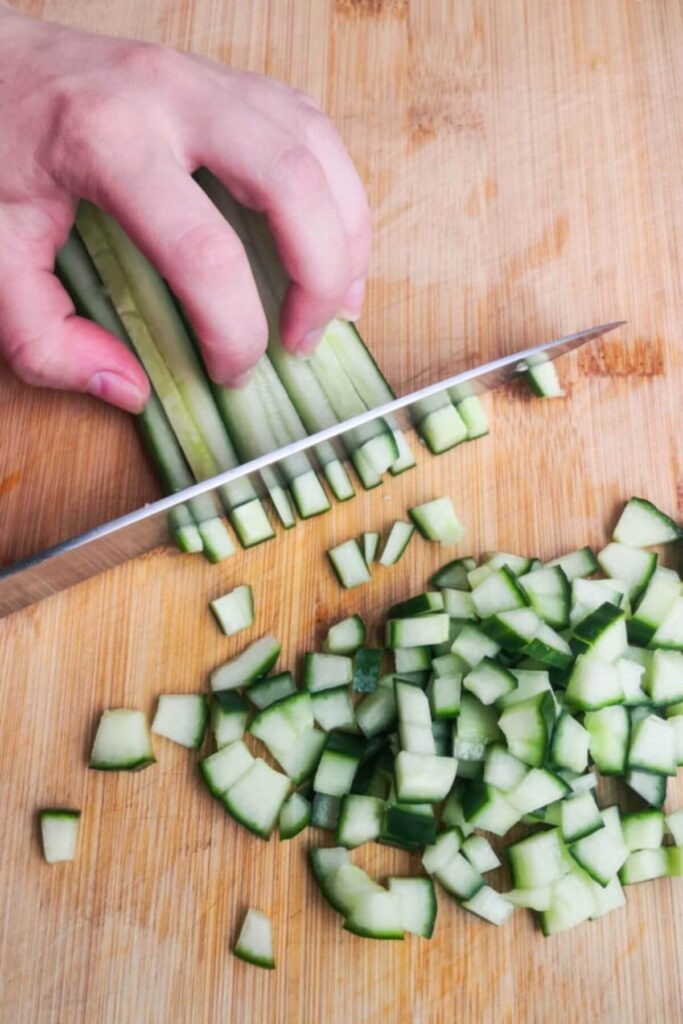 Chopping cucumber strips into small cubes with a knife on a wooden board.