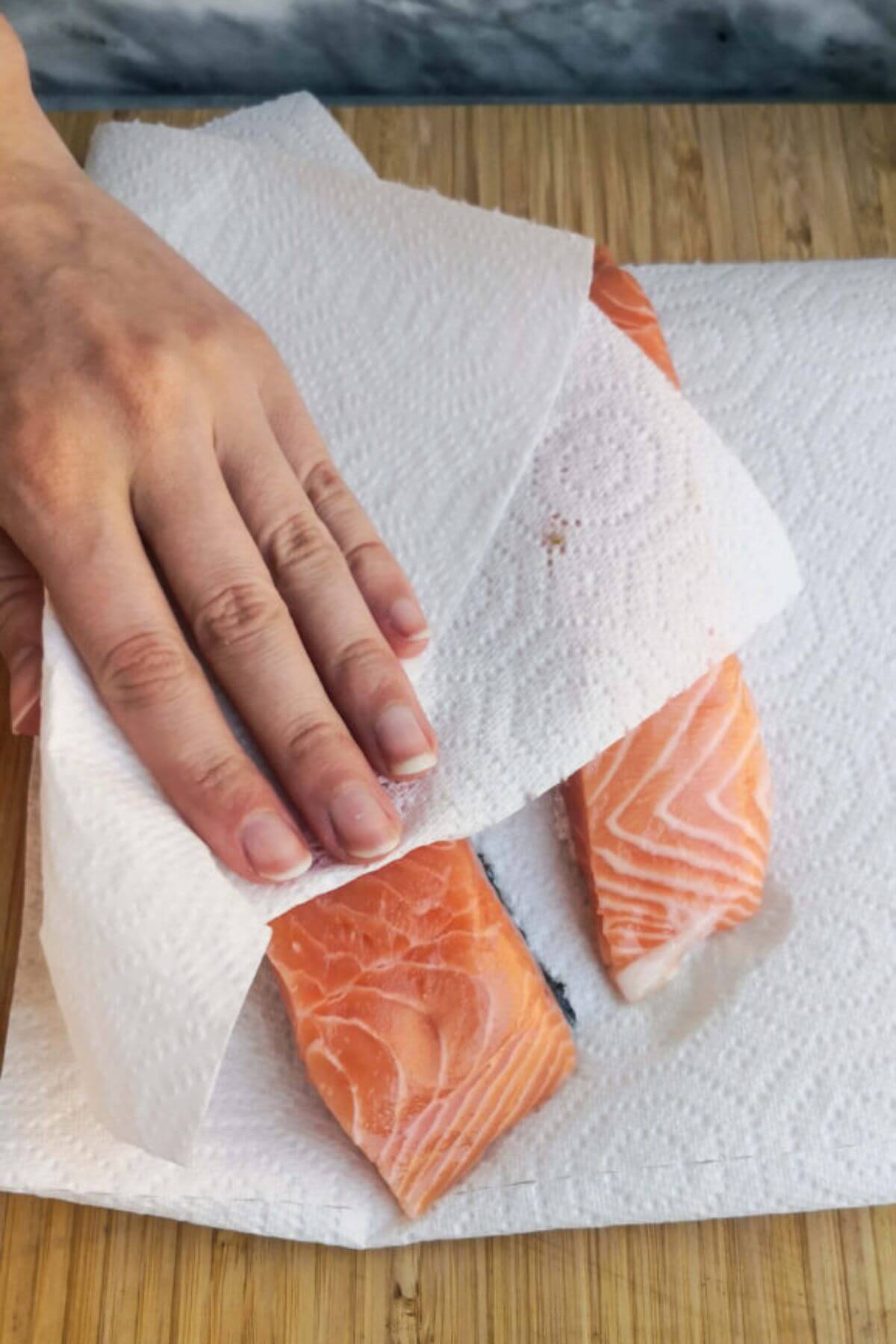 Hands and a paper towel patting dry two salmon fillets on a wooden board.