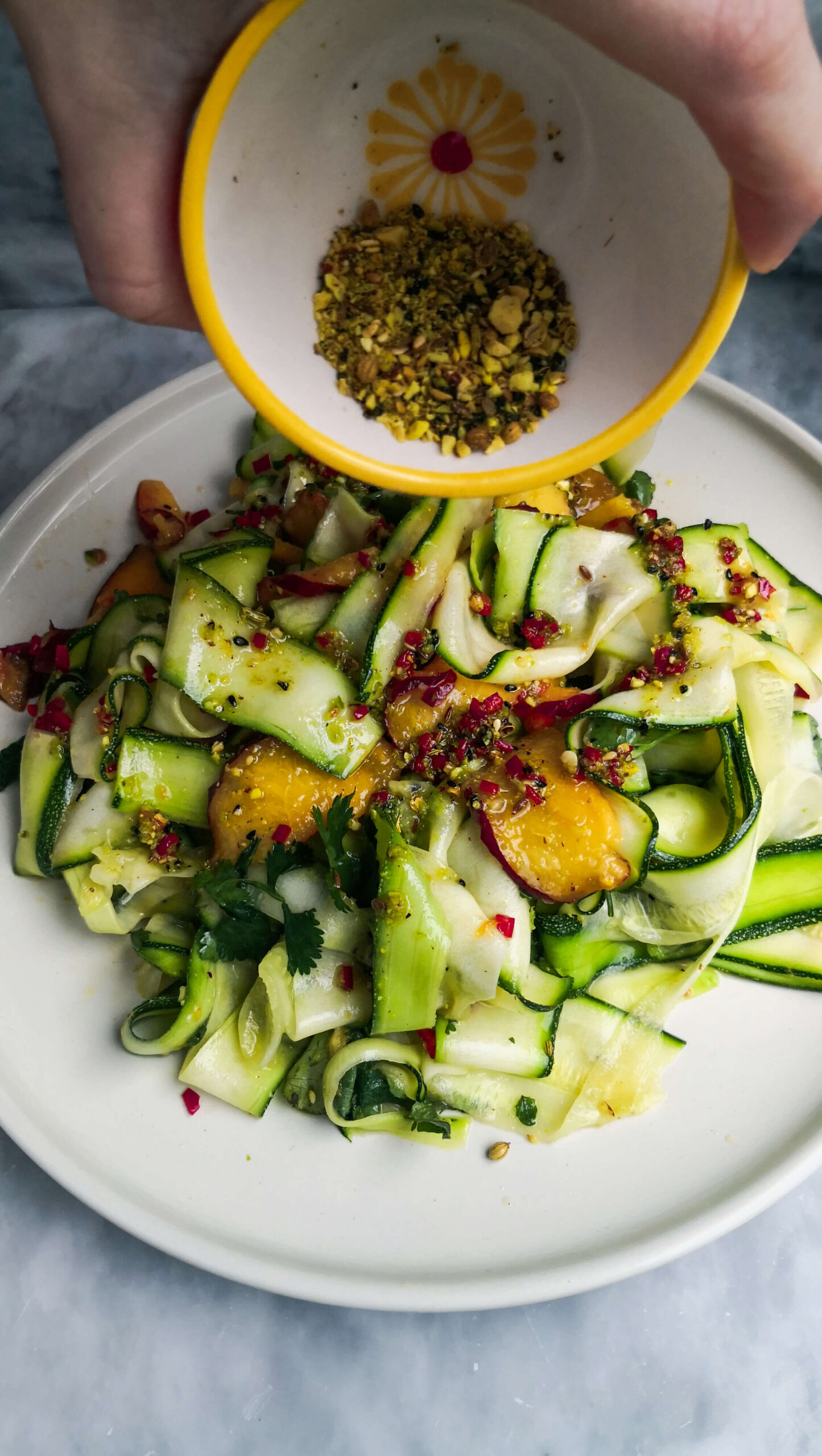 Dukkah being added to raw courgette and peach salad on a white plate.