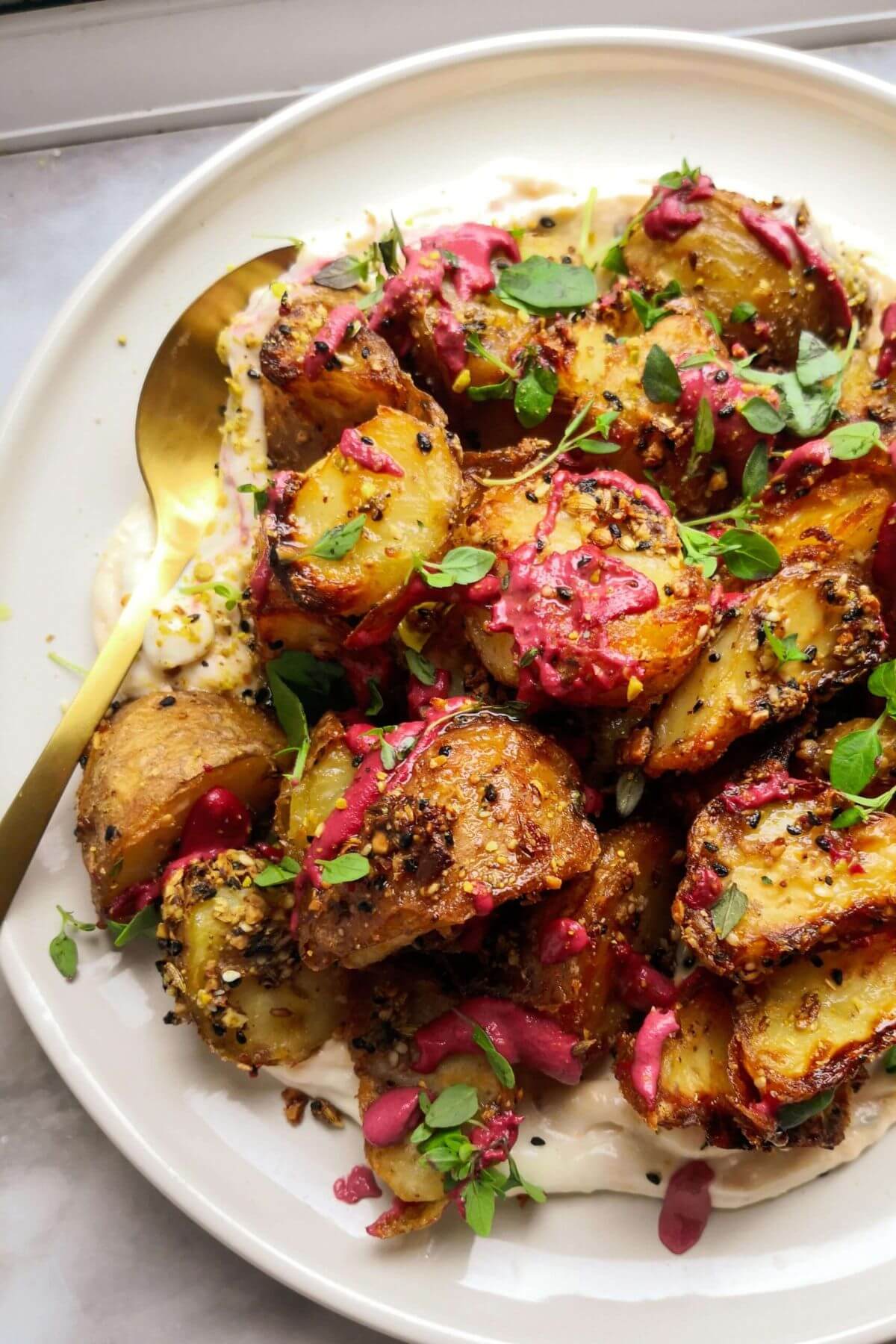 Crispy dukkah potatoes on a white plate with a gold spoon, with thyme and pink tahini on top.