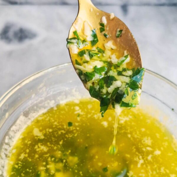 Garlic butter sauce in a small glass bowl, with a gold spoon with sauce on it, on a grey marble background.