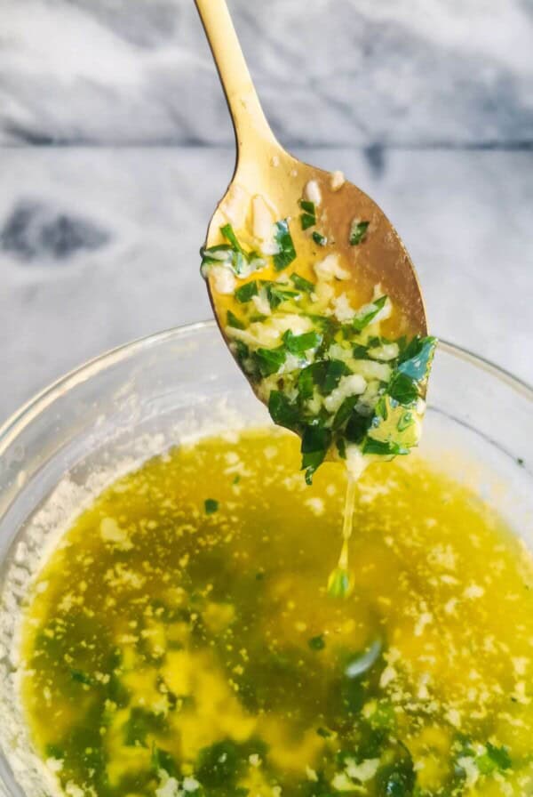 Garlic butter sauce in a small glass bowl, with a gold spoon with sauce on it, on a grey marble background.