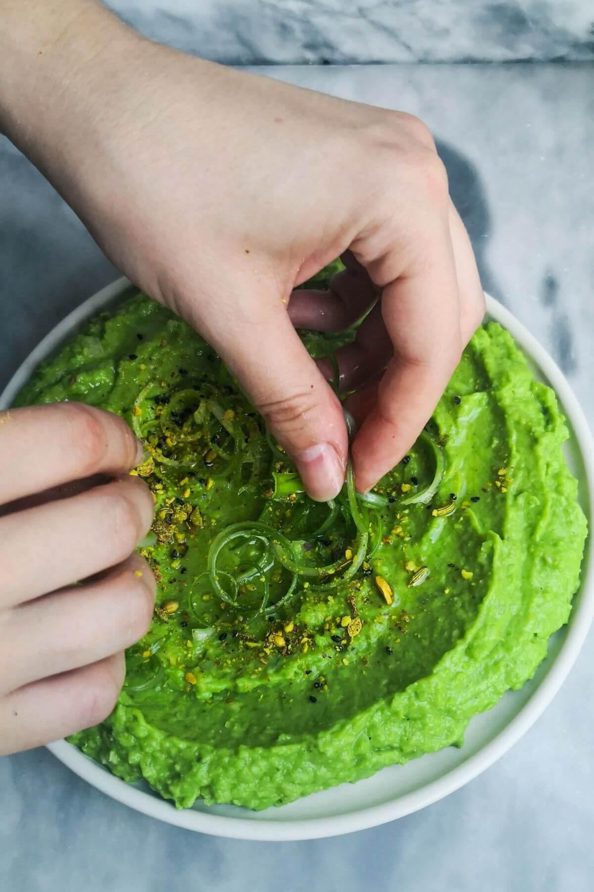 Hands adding curly spring onions to the plate of pea hummus on a grey marble background.