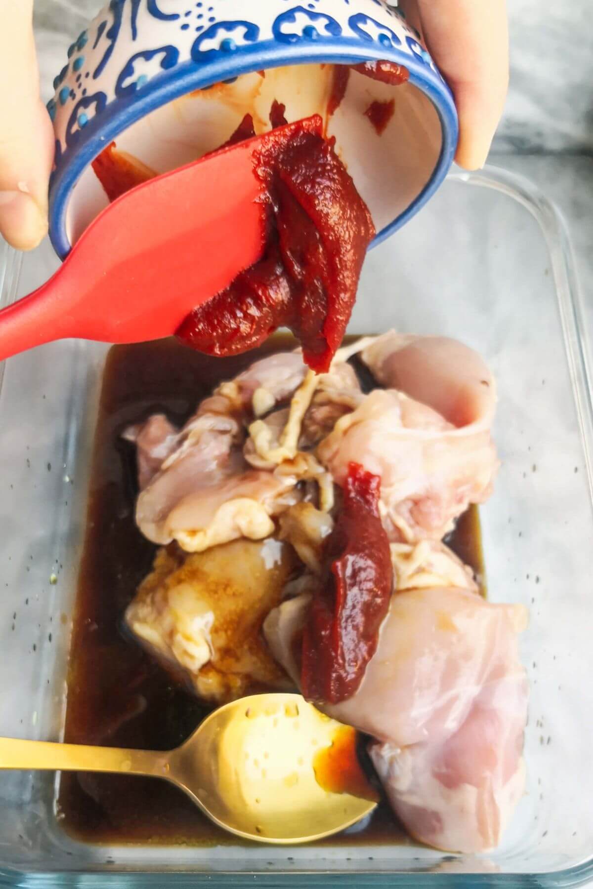Gochujang paste being added to chicken in a large glass container.