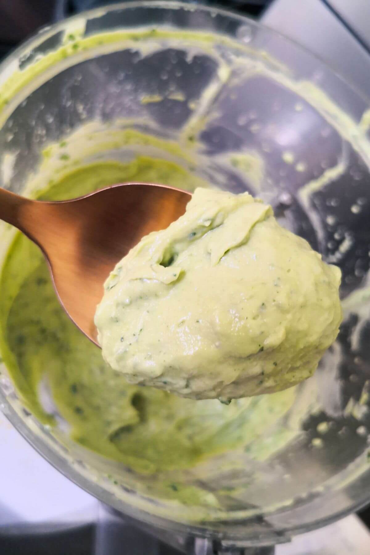 Close up of spoon with green tahini on it, with food processor bowl in the background.
