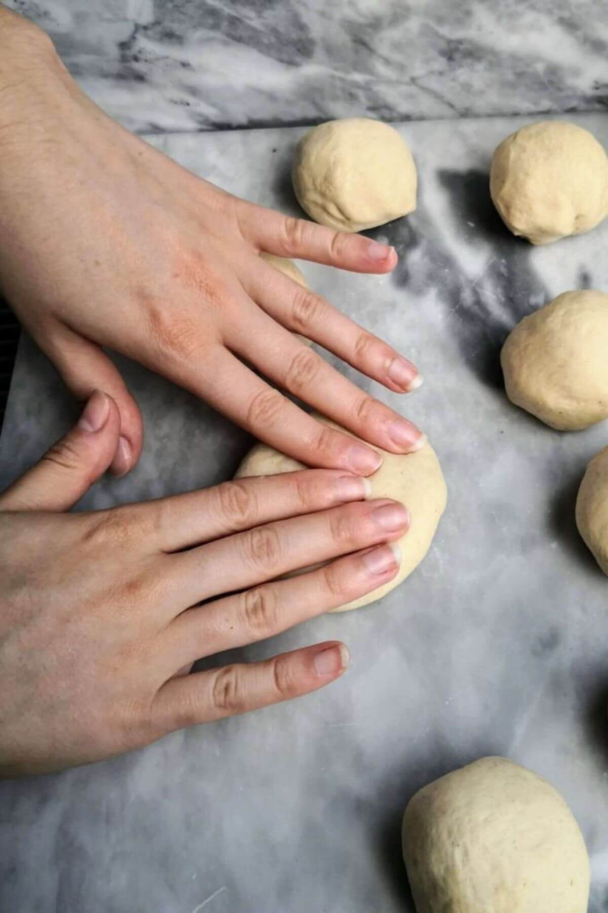 Hands lightly pressing down dough ball into an oval on a grey marble background.