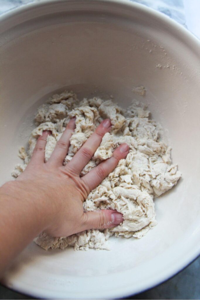 Hand pressing down into bao dough in a large silver mixing bowl.