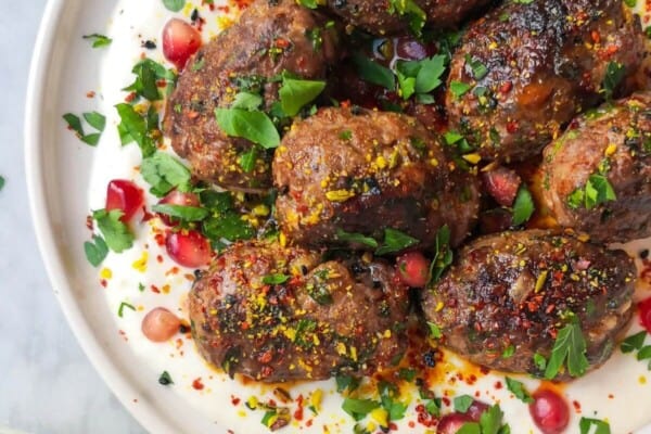 Lamb kofta with parsley, pomegranate seeds and dukkah on a white plate with yogurt tahini sauce, with a bowl of yogurt tahini sauce on the side.