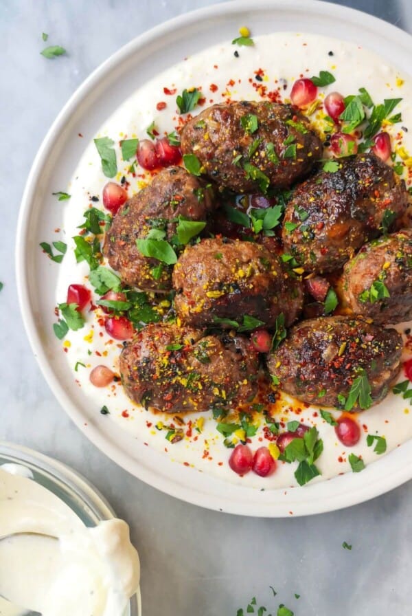 Lamb kofta with parsley, pomegranate seeds and dukkah on a white plate with yogurt tahini sauce, with a bowl of yogurt tahini sauce on the side.