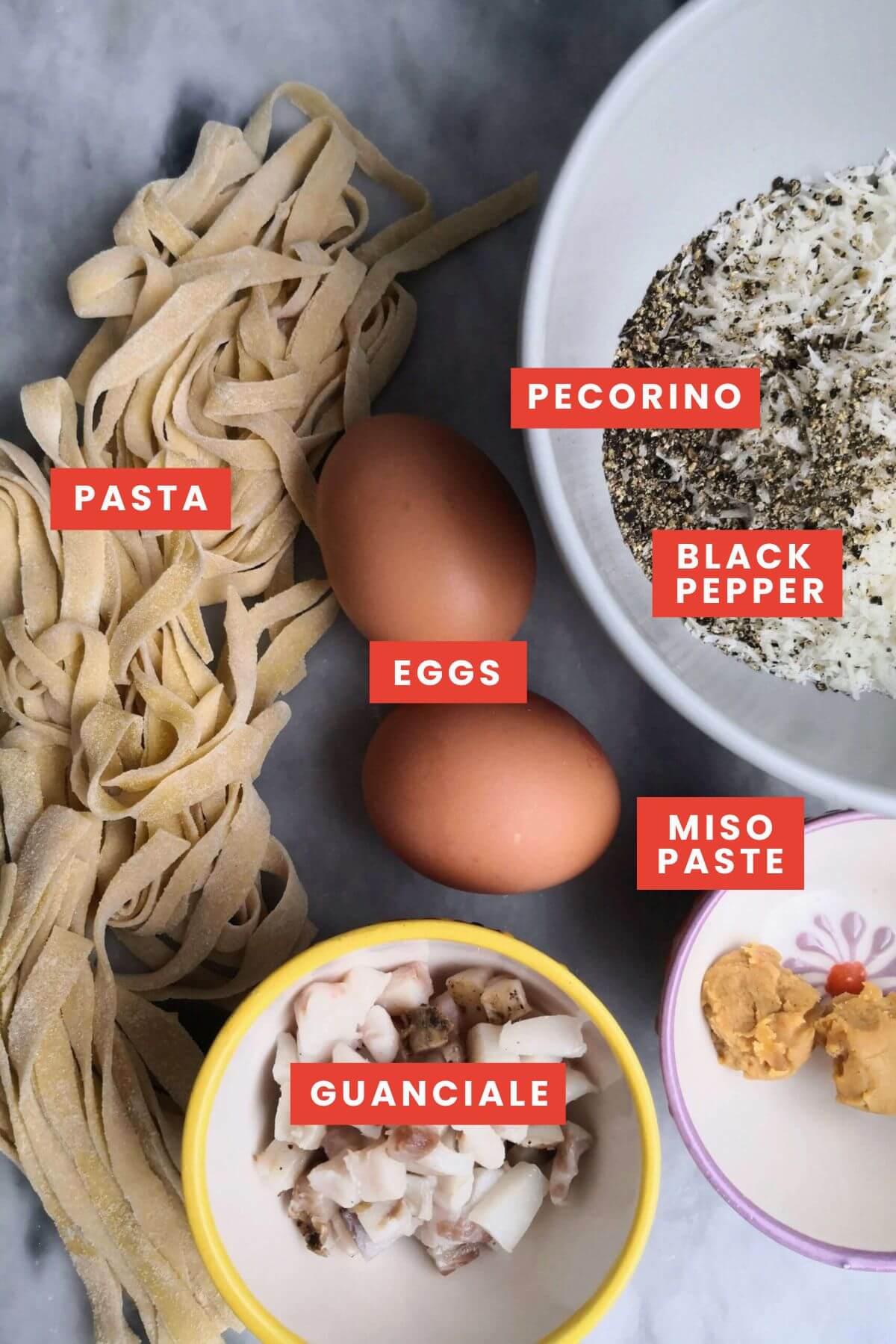 Ingredients for miso carbonara laid out on a grey marble board - fettuccine, eggs, pecorino romano, black pepper, guanciale and miso paste.