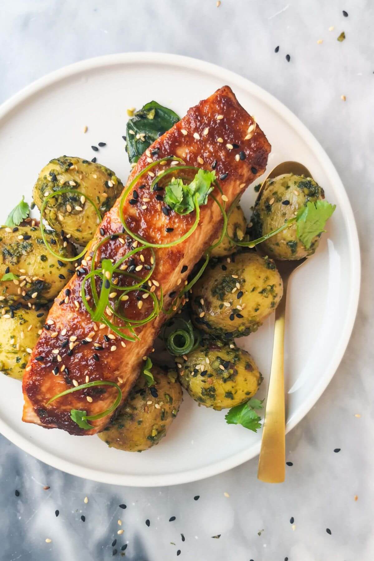 Miso glazed salmon on top of herby potatoes on a white plate, with sesame seeds, coriander and spring onion on top.