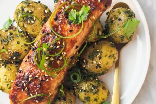 Miso glazed salmon on top of herby potatoes on a white plate, with sesame seeds, coriander and spring onion on top.