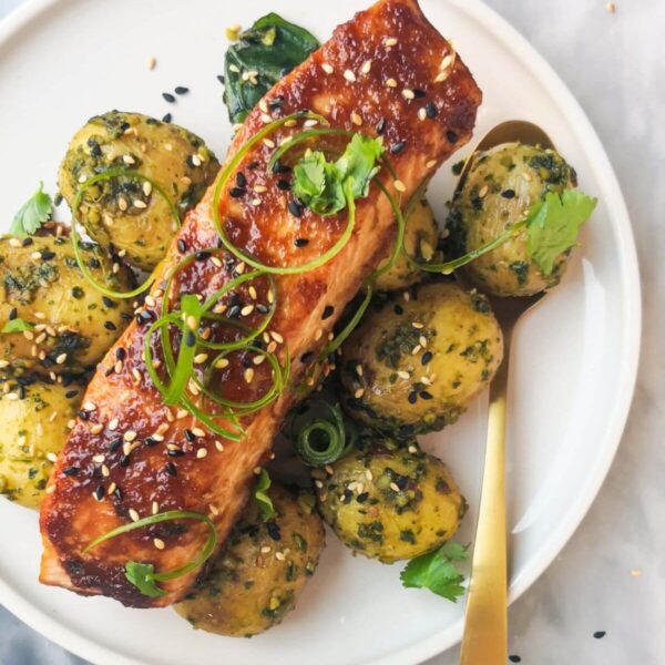 Miso glazed salmon on top of herby potatoes on a white plate, with sesame seeds, coriander and spring onion on top.