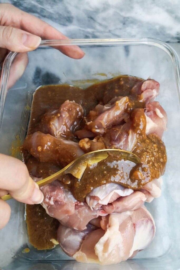 Gold spoon mixing chicken thighs with the miso marinade in a glass container on a grey marble background.