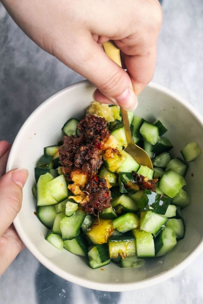 Hand holding a small gold spoon mixing the cucumber salad in a small white bowl.