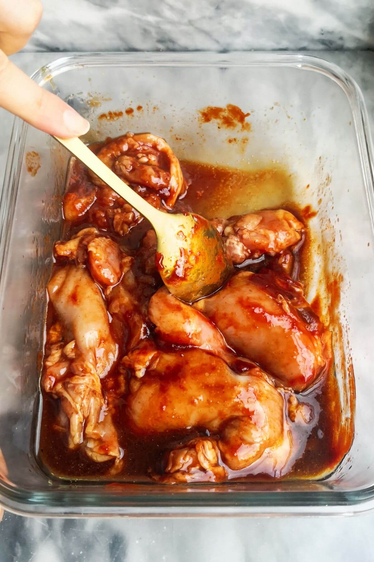 Gold spoon mixing chicken thighs through gochujang marinade in a glass container.