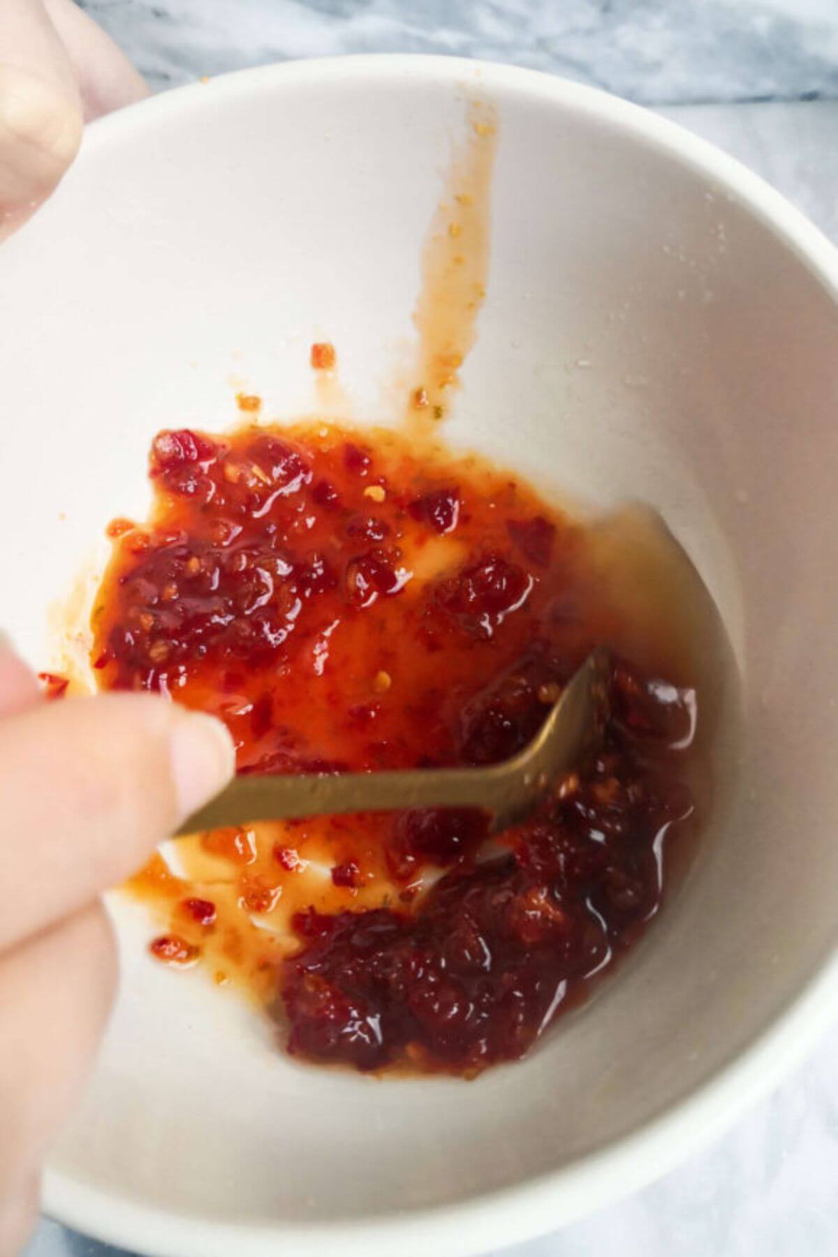 A hand holding a gold spoon mixing chilli jam and lime juice in a small white bowl.