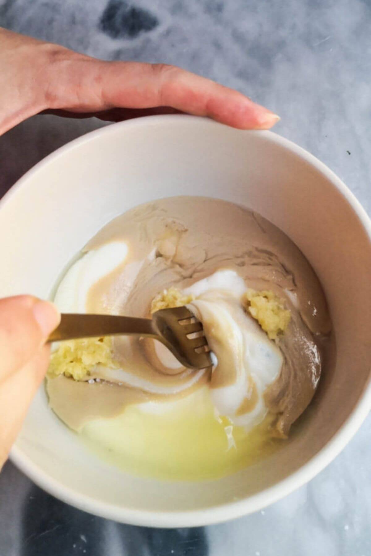 Yogurt tahini sauce ingredients being mixed together with a gold fork in a small white bowl.