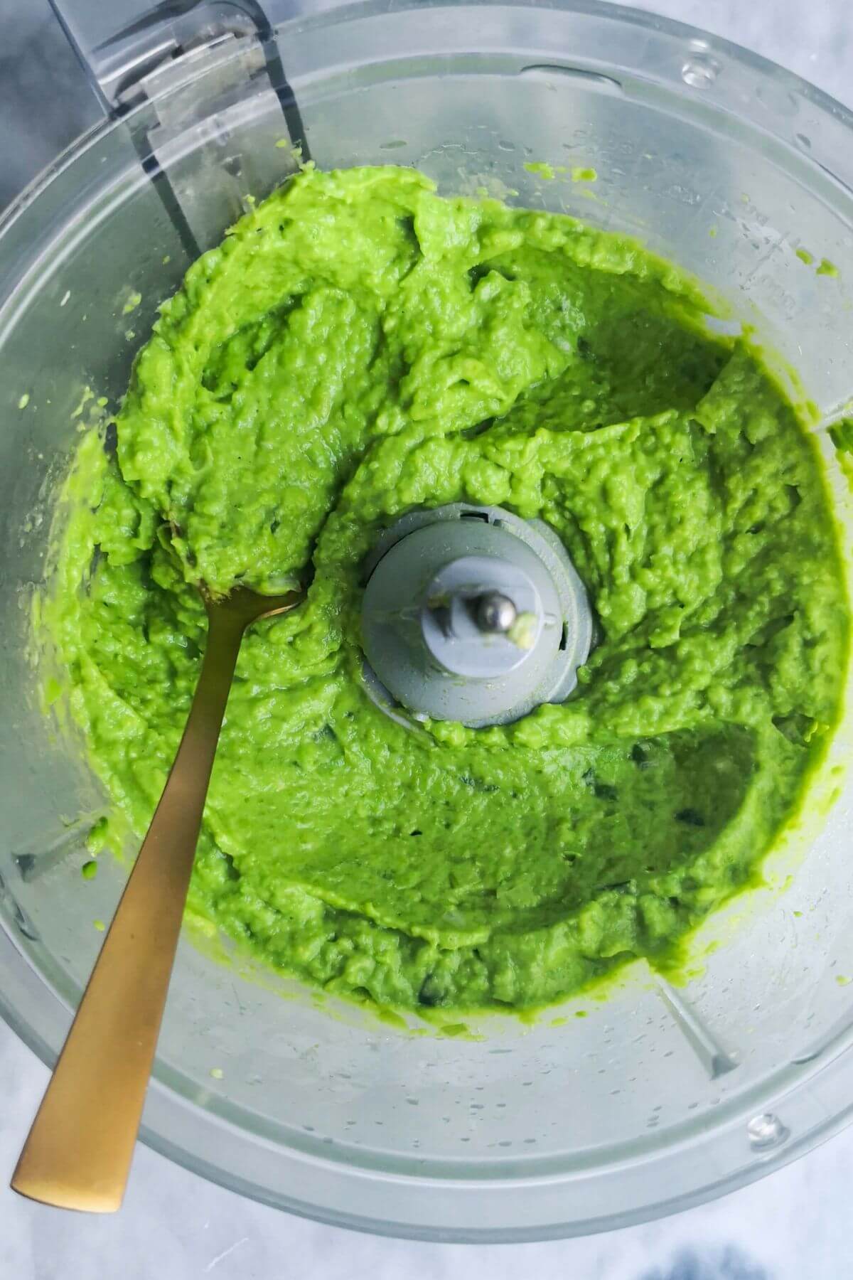A gold spoon in the bowl of a food processor with blitzed pea hummus.