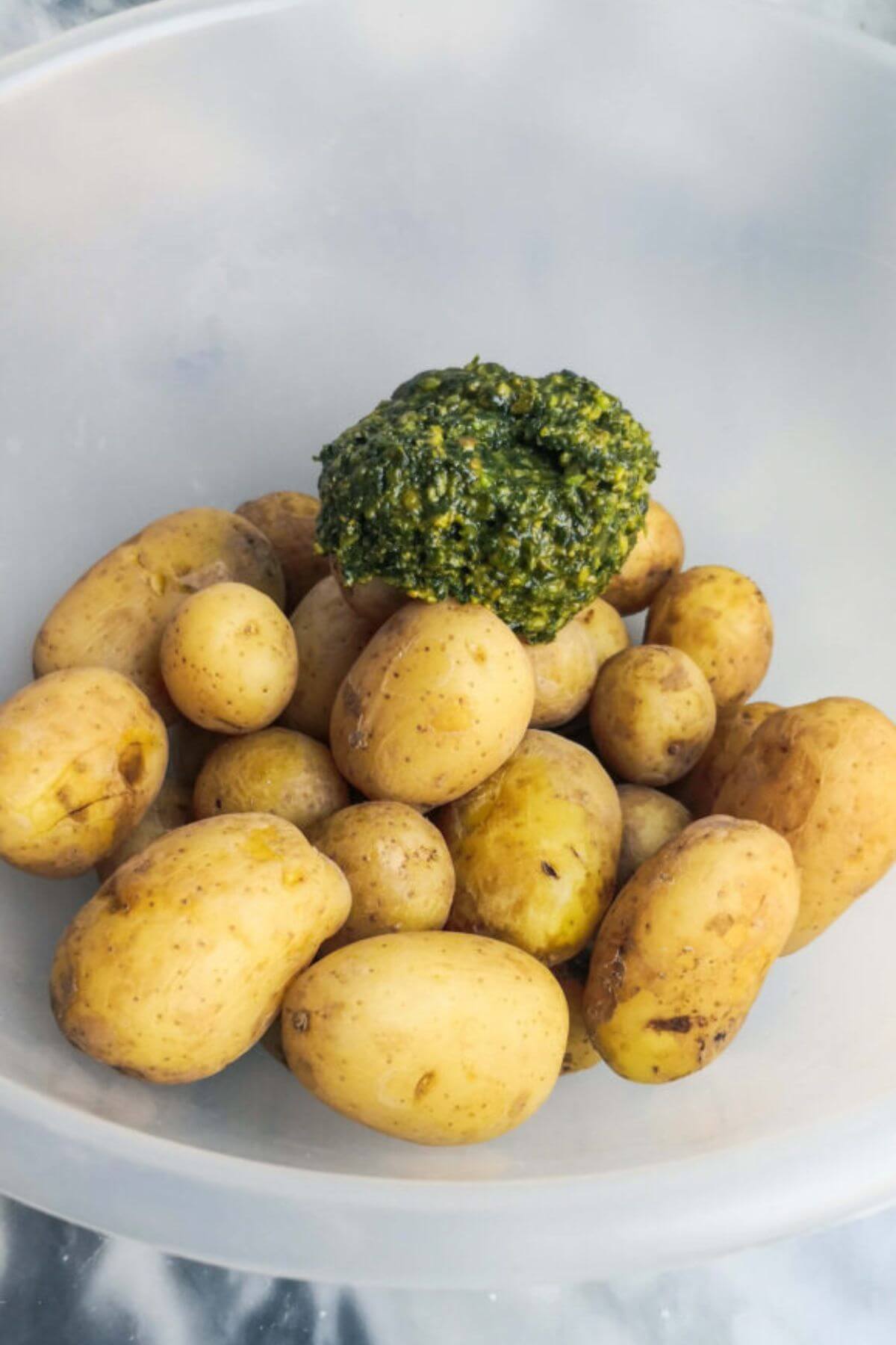 Pesto added to a large clear mixing bowl with potatoes in it.