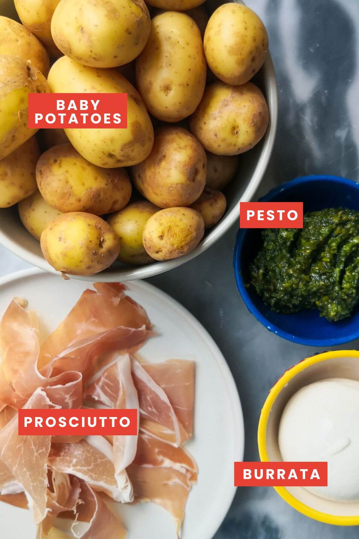 Ingredients for pesto potatoes with crispy prosciutto and burrata laid out on a grey marble background.