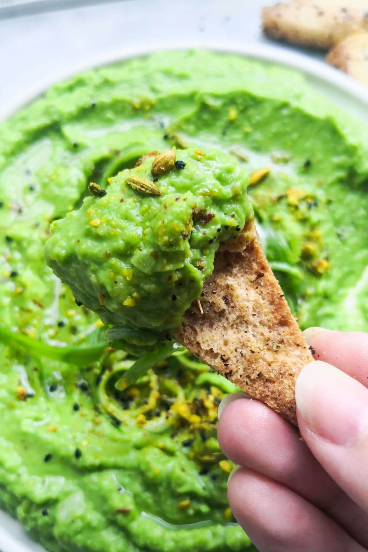Close up of a pita chip with pea hummus on it, with a plate of pea hummus in the background.