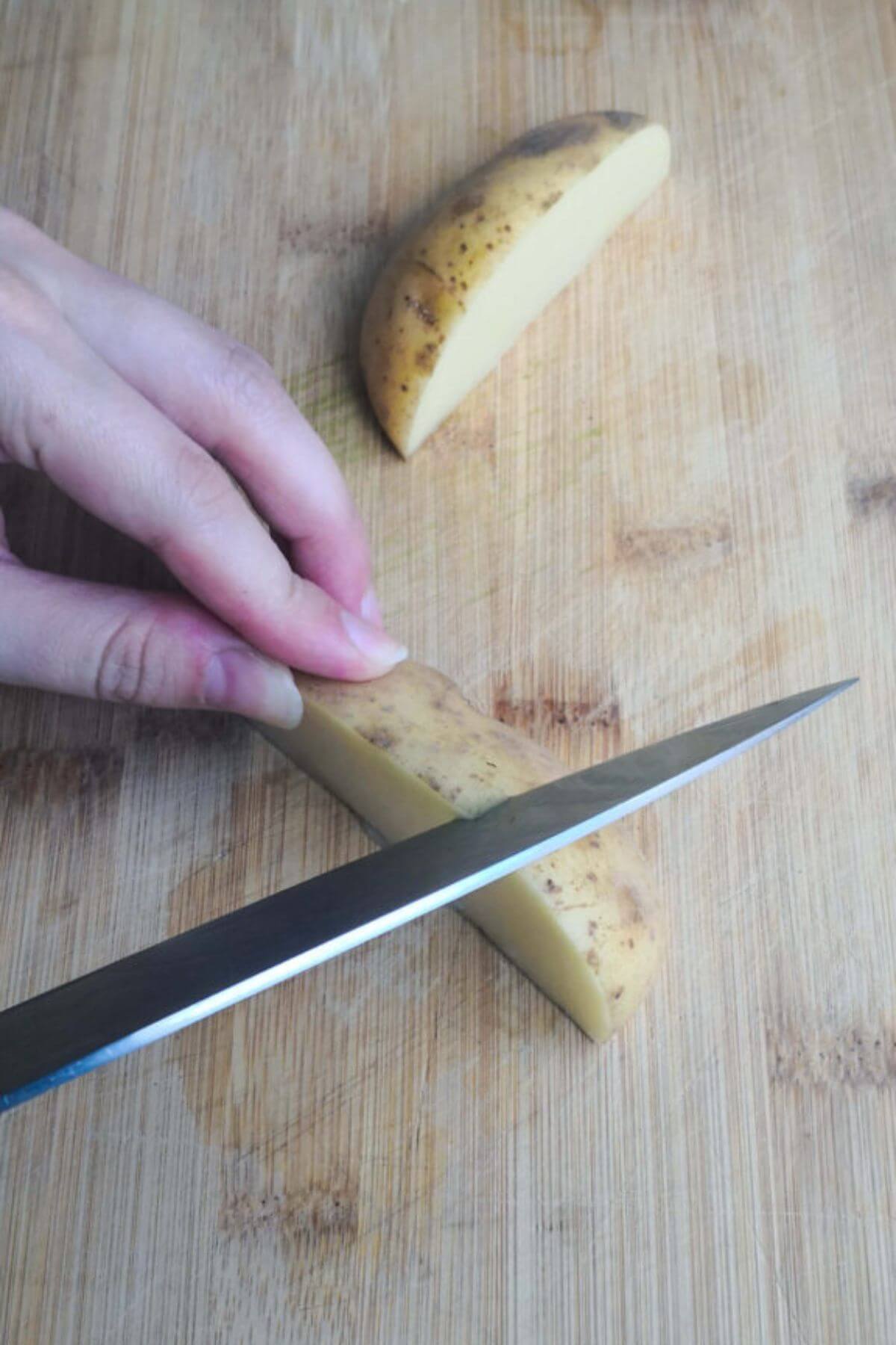 Hand and knife chopping a piece of potato into 2 chunks on a wooden board.