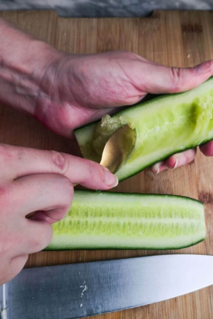 Small gold spoon scooping the seeds out of a cucumber on a wooden board.
