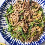 Sesame soba noodles with cucumber and broccolini in a white and blue bowl with chopsticks, with a small bowl of spring onions on the side.