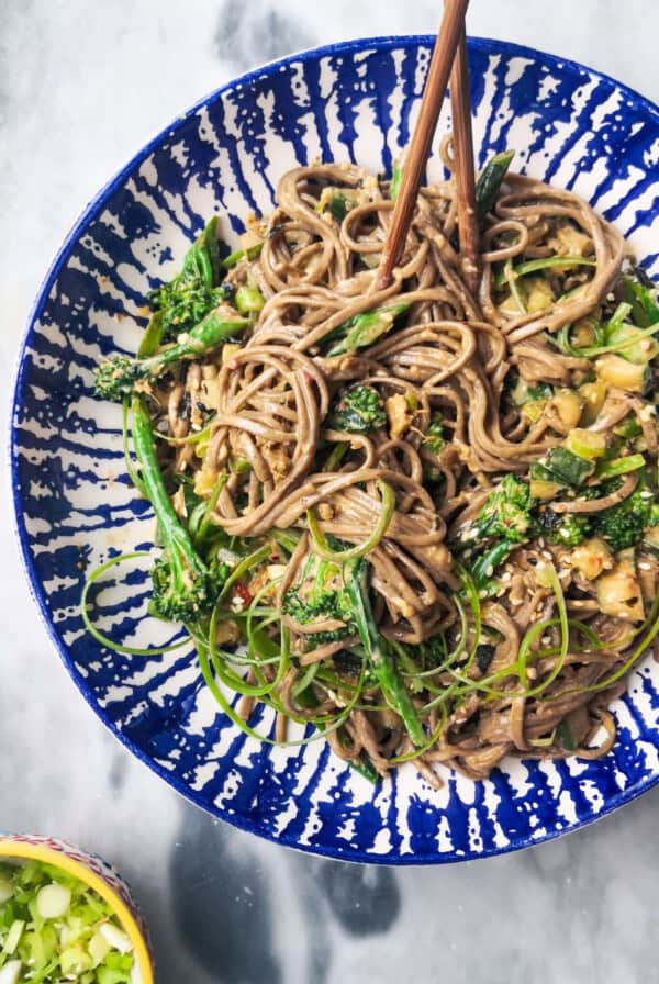 Sesame soba noodles with cucumber and broccolini in a white and blue bowl with chopsticks, with a small bowl of spring onions on the side.