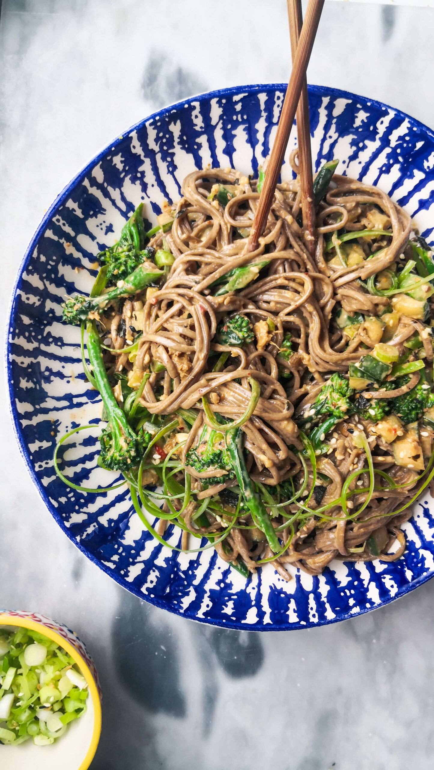 Sesame soba noodles with cucumber and broccolini in a white and blue bowl with chopsticks, with a small bowl of spring onions on the side.