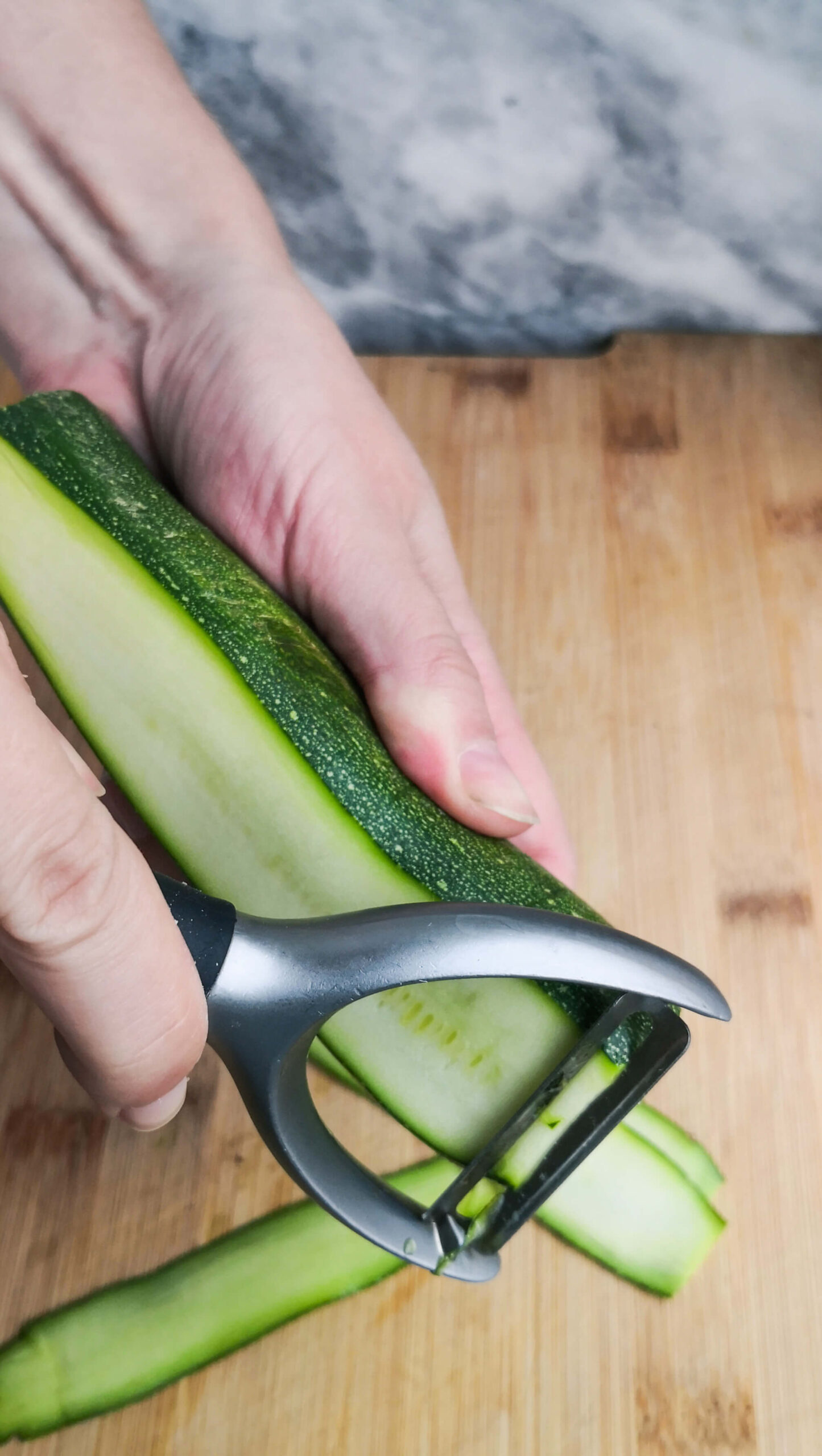 Hand holding a courgette and starting to peel a strip off with a vegetable peeler.